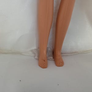 K&ouml;nnte beinhalten: Nahaufnahme der Beine und F&uuml;&szlig;e einer Puppe. Die Beine der Puppe sind hellbraun, und die F&uuml;&szlig;e weisen einige Abnutzungserscheinungen auf. Die Puppe tr&auml;gt ein wei&szlig;es Kleid, dessen Saum am unteren Bildrand sichtbar ist.