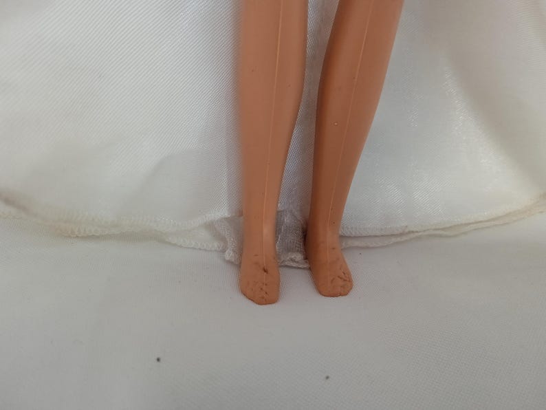 K&ouml;nnte beinhalten: Nahaufnahme der Beine und F&uuml;&szlig;e einer Puppe. Die Beine der Puppe sind hellbraun, und die F&uuml;&szlig;e weisen einige Abnutzungserscheinungen auf. Die Puppe tr&auml;gt ein wei&szlig;es Kleid, dessen Saum am unteren Bildrand sichtbar ist.