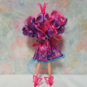 K&ouml;nnte beinhalten: Eine Puppe mit rosa und lila Haaren, die ein rosa Kleid mit blauer Bord&uuml;re tr&auml;gt. Die Puppe hat rosa Schuhe und eine rosa Krone. Das Kleid hat ein Muster aus Sternen und Schleifen. Die Puppe steht auf einer wei&szlig;en Oberfl&auml;che.