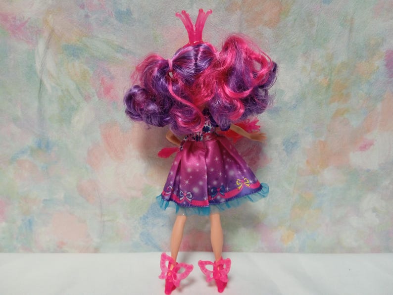 K&ouml;nnte beinhalten: Eine Puppe mit rosa und lila Haaren, die ein rosa Kleid mit blauer Bord&uuml;re tr&auml;gt. Die Puppe hat rosa Schuhe und eine rosa Krone. Das Kleid hat ein Muster aus Sternen und Schleifen. Die Puppe steht auf einer wei&szlig;en Oberfl&auml;che.