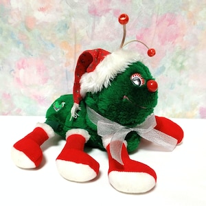 K&ouml;nnte beinhalten: Ein gr&uuml;ner Pl&uuml;sch-Weihnachtsk&auml;fer, der einen roten und wei&szlig;en Weihnachtsmann-Hut mit wei&szlig;em Pelzbesatz tr&auml;gt. Der K&auml;fer hat rote und wei&szlig;e Stiefel und ein wei&szlig;es Band um den K&ouml;rper.