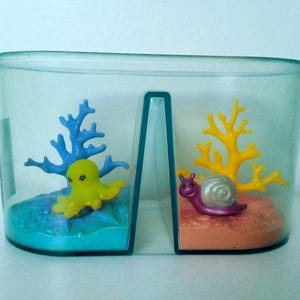 Puede incluir: Un recipiente de plástico transparente con dos compartimentos, cada uno con una escena submarina en miniatura. Un lado presenta un pulpo amarillo con coral azul, el otro un caracol morado con coral amarillo. La base de cada escena es de diferente color.