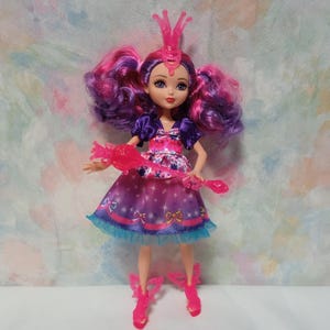 K&ouml;nnte beinhalten: Eine Modepuppe mit lila und rosa Haaren, die ein lila und rosa Kleid mit blauem Besatz tr&auml;gt. Die Puppe hat eine rosa Krone und passende Schuhe. Sie h&auml;lt einen rosa Zauberstab.