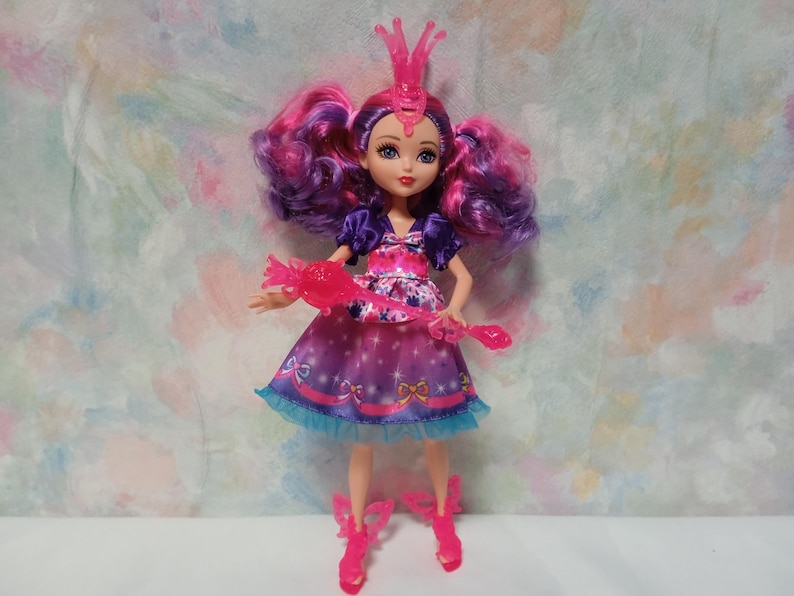 K&ouml;nnte beinhalten: Eine Modepuppe mit lila und rosa Haaren, die ein lila und rosa Kleid mit blauem Besatz tr&auml;gt. Die Puppe hat eine rosa Krone und passende Schuhe. Sie h&auml;lt einen rosa Zauberstab.