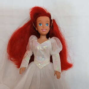K&ouml;nnte beinhalten: Puppe einer Figur mit langen, leuchtend roten Haaren und blauen Augen, die ein wei&szlig;es Hochzeitskleid mit schillernden Akzenten tr&auml;gt. Das Kleid hat lange &Auml;rmel und einen mehrlagigen Rock. Ein wei&szlig;er Schleier ist am Haar der Puppe befestigt.