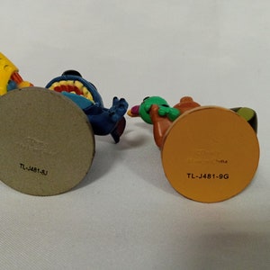 Puede incluir: Dos peque&ntilde;as figuras de pl&aacute;stico, una azul con una pistola de agua amarilla y la otra verde y marr&oacute;n con una pistola de agua amarilla y negra. Las bases de las figuras son marrones y naranjas y tienen el texto "TL-J481-BJ" y "TL-J481-9G" impreso en ellas.