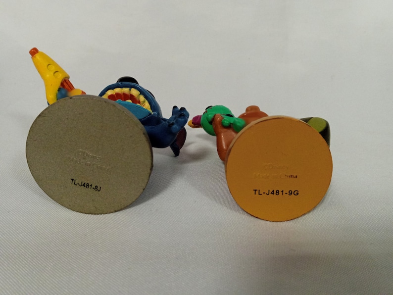 Puede incluir: Dos peque&ntilde;as figuras de pl&aacute;stico, una azul con una pistola de agua amarilla y la otra verde y marr&oacute;n con una pistola de agua amarilla y negra. Las bases de las figuras son marrones y naranjas y tienen el texto "TL-J481-BJ" y "TL-J481-9G" impreso en ellas.