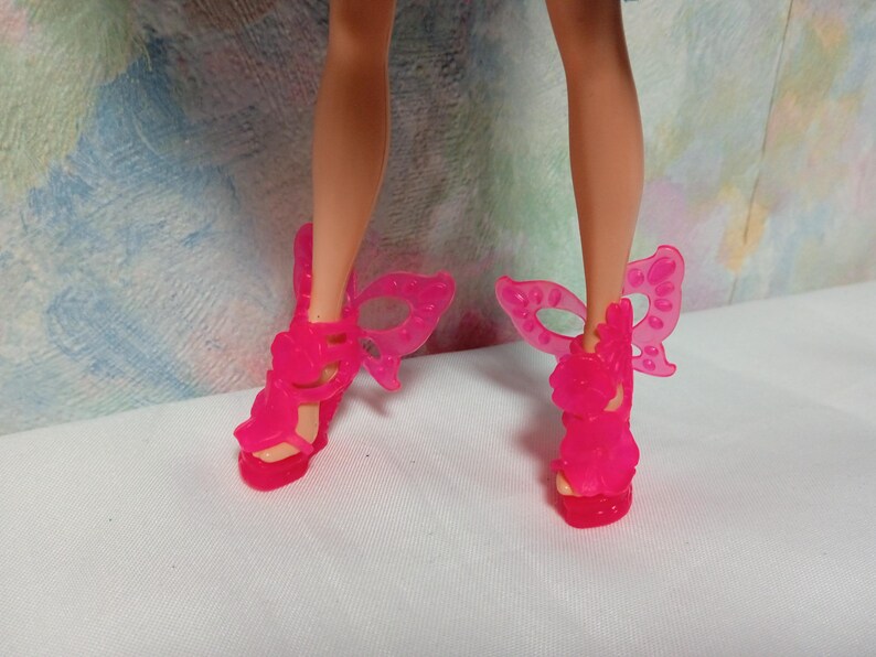 K&ouml;nnte beinhalten: Ein Paar leuchtend pinkfarbene Puppenschuhe. Die Schuhe haben einen hohen Absatz und ein Schmetterlingsfl&uuml;gel-Design an der Seite. Die Schuhe sind aus durchscheinendem Kunststoff.