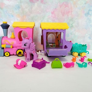 May include: A pink and purple toy train set with a purple pony figure, a pink engine car, a purple passenger car, and a green and yellow caboose. The set includes various accessories, such as a pink hat, a purple purse, and a green purse.