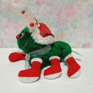 K&ouml;nnte beinhalten: Ein gr&uuml;ner Pl&uuml;sch-Weihnachtsk&auml;fer mit roter und wei&szlig;er Weihnachtsmannm&uuml;tze, roten Stiefeln und einem wei&szlig;en Band.