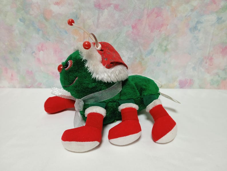 K&ouml;nnte beinhalten: Ein gr&uuml;ner Pl&uuml;sch-Weihnachtsk&auml;fer mit roter und wei&szlig;er Weihnachtsmannm&uuml;tze, roten Stiefeln und einem wei&szlig;en Band.