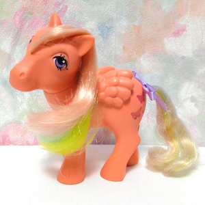 Op de afbeelding: Een vintage oranje speelgoedpony met een vloeiende manen en staart in roze, gele en groene tinten. De pony heeft vlindervleugels en een paarse strik. Het speelgoed is een verzamelobject.
