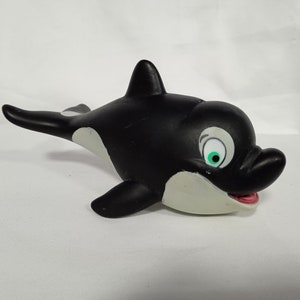 K&ouml;nnte beinhalten: Schwarzes und wei&szlig;es Spielzeug-Orca aus Gummi mit einem gr&uuml;nen Auge und einem rosa Mund. Der Orca schwimmt nach rechts.