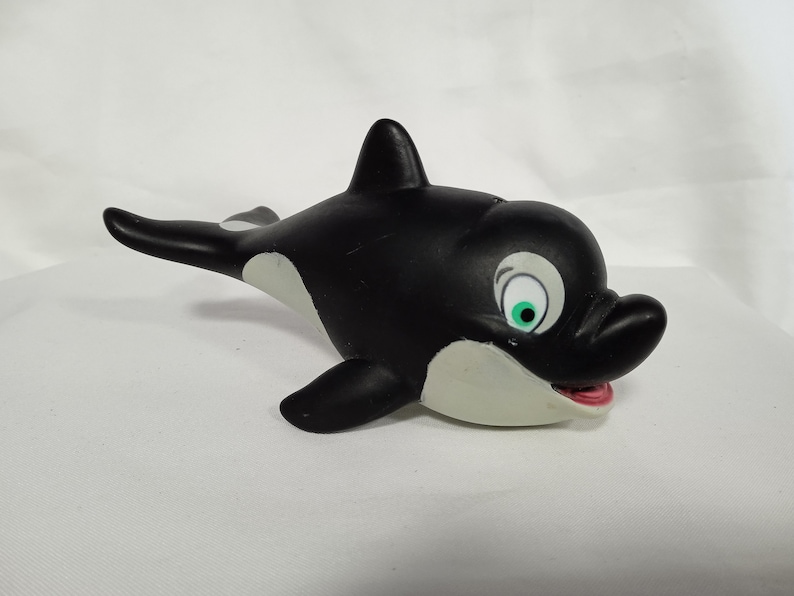 K&ouml;nnte beinhalten: Schwarzes und wei&szlig;es Spielzeug-Orca aus Gummi mit einem gr&uuml;nen Auge und einem rosa Mund. Der Orca schwimmt nach rechts.