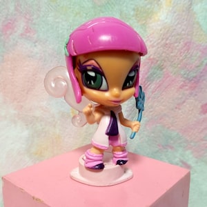 Peut inclure: Petite figurine de fée de dessin animé avec un casque rose, une robe blanche et des accents violets. La fée a les yeux verts et tient une baguette bleue. La figurine est sur une base carrée rose.