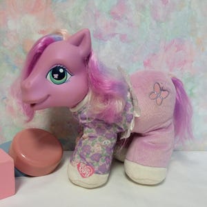 Könnte beinhalten: Ein rosa Plüschpony, das einen lila-weißen Blumenpyjama mit einem Schmetterling auf dem Rücken trägt. Das Pony hat eine rosa-weiße Mähne und einen Schwanz.