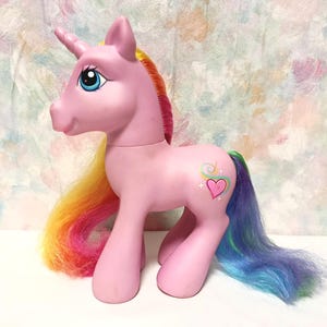 Puede incluir: Un juguete de unicornio rosa con un cuerno moldeado, ojos azules y una larga melena y cola de pelo sintético con los colores del arcoíris. El unicornio tiene un símbolo en forma de corazón con un diseño de remolino colorido en su flanco izquierdo.