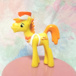 Peut inclure: Figurine de poney jouet jaune avec une crinière et une queue orange, portant un tablier blanc et un chapeau de chef. Le poney a une expression souriante et se tient sur une surface rose.