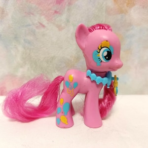 Può includere: Una statuina di pony giocattolo rosa con criniera e coda rosa. Il pony ha accenti blu e gialli, tra cui palloncini sul lato e una collana blu. Il pony ha grandi occhi blu e un disegno giallo e blu attorno a un occhio.