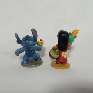 Puede incluir: Dos peque&ntilde;as figuras de pl&aacute;stico, una Stitch azul de Lilo & Stitch sosteniendo un objeto amarillo, y la otra una ni&ntilde;a de cabello casta&ntilde;o con un vestido rojo sosteniendo un objeto verde y amarillo.