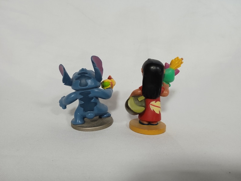Puede incluir: Dos peque&ntilde;as figuras de pl&aacute;stico, una Stitch azul de Lilo & Stitch sosteniendo un objeto amarillo, y la otra una ni&ntilde;a de cabello casta&ntilde;o con un vestido rojo sosteniendo un objeto verde y amarillo.