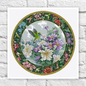 May include: A cross-stitch design depicting a hummingbird hovering near white, purple, and blue petunias. The circular design is framed by a border of flowers and hummingbirds, with a gold-coloured outer ring. The design is on a white background.