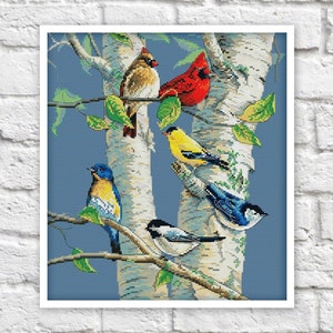 May include: A cross-stitch pattern featuring a birch tree with six birds perched on its branches. The birds are a cardinal, a blue jay, a chickadee, a goldfinch, and two other small birds. The background is a light blue color.