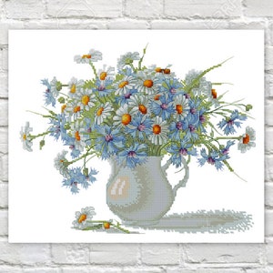 May include: A cross-stitch depiction of a bouquet of daisies and cornflowers in a white pitcher. The flowers are in shades of blue, white, and yellow, with green stems. The pitcher casts a shadow on the white surface.