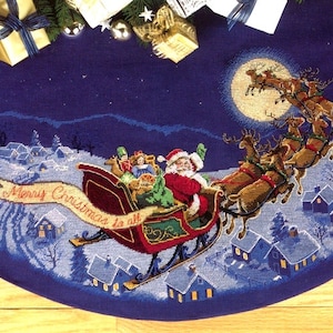 Peut inclure: Une jupe de sapin de Noël bleue avec une scène de village enneigé et le Père Noël volant dans son traîneau tiré par des rennes. Le texte "Merry Christmas to all" est sur une bannière dans le traîneau.