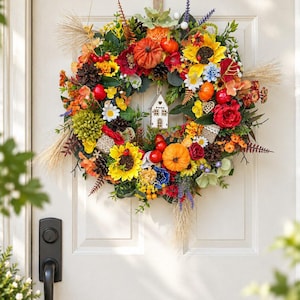 Könnte beinhalten: Ein runder Kranz mit einer Mischung aus künstlichen Sonnenblumen, orangefarbenen und roten Kürbissen, Tannenzapfen, roten Beeren und grünem Laub. Ein kleines weißes Holzhaus hängt in der Mitte, umgeben von getrockneten Weizenhalmen.