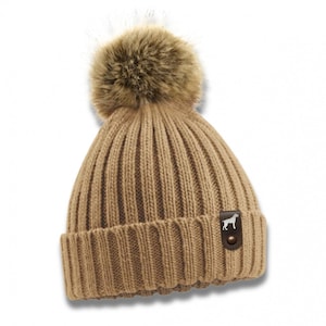 Könnte beinhalten: Eine braune Strickmütze mit einem Kunstfellbommel oben. Die Mütze hat einen kleinen braunen Lederfleck mit einer weißen Silhouette eines Hundes.