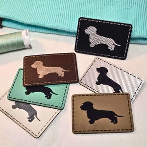 Könnte beinhalten: Sechs verschiedene Dackel-Patches in verschiedenen Farben und Materialien. Die Patches sind alle gleich groß und haben die gleiche Form und zeigen eine Silhouette eines Dackels. Die Patches sind perfekt, um Ihren Kleidungsstücken, Taschen oder anderem Zubehör eine persönliche Note zu verleihen.