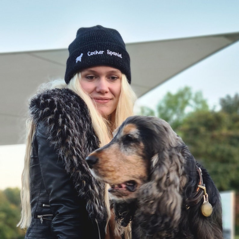 Op de afbeelding: Zwarte gebreide beanie met de tekst "Cocker Spaniel" en een wit honden silhouet. De beanie wordt gedragen door een persoon met blond haar. Een Cocker Spaniel hond is ook zichtbaar in de afbeelding.