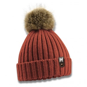 Könnte beinhalten: Eine rostfarbene Strickmütze mit einem braunen Kunstfellbommel oben. Die Mütze hat einen kleinen braunen Lederfleck mit einer weißen Silhouette eines Hundes.