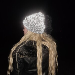 Op de afbeelding: Een reflecterende beanie-muts met een gespikkeld patroon, gedragen door een persoon met lang blond haar. De hoed lijkt helder op te lichten tegen de donkere achtergrond. De persoon draagt een zwarte leren jas met een imitatiebontkraag.