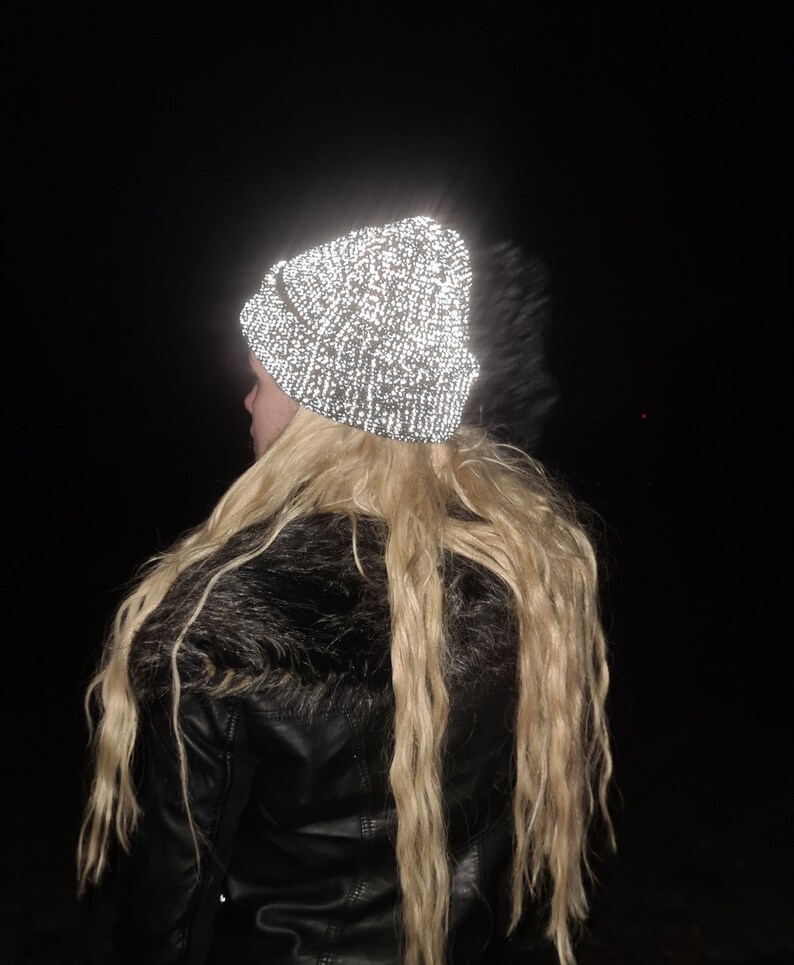 Op de afbeelding: Een reflecterende beanie-muts met een gespikkeld patroon, gedragen door een persoon met lang blond haar. De hoed lijkt helder op te lichten tegen de donkere achtergrond. De persoon draagt een zwarte leren jas met een imitatiebontkraag.