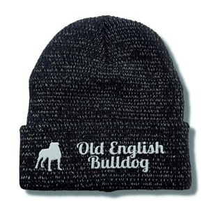 Op de afbeelding: Zwart-witte gebreide muts met de woorden "Old English Bulldog" in het wit geborduurd. De muts heeft een silhouet van een Old English Bulldog. Een klassiek, warm winteraccessoire.