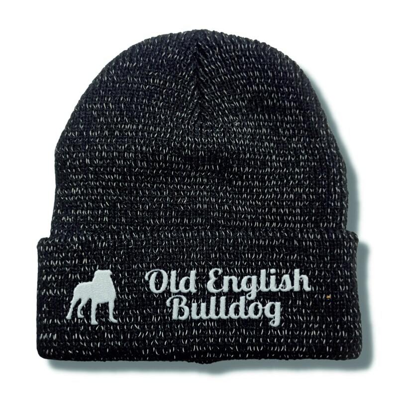 Op de afbeelding: Zwart-witte gebreide muts met de woorden "Old English Bulldog" in het wit geborduurd. De muts heeft een silhouet van een Old English Bulldog. Een klassiek, warm winteraccessoire.