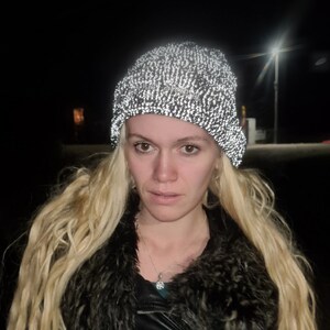 Op de afbeelding: Een reflecterende gebreide beanie-muts in een zwart-wit gespikkeld patroon. De hoed wordt gedragen door een persoon met lang blond haar, een zwarte jas en een bontkraag. De achtergrond is donker met een straatlantaarn.