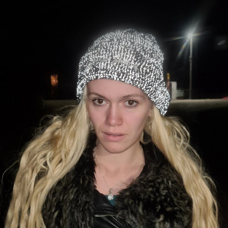 Op de afbeelding: Een reflecterende gebreide beanie-muts in een zwart-wit gespikkeld patroon. De hoed wordt gedragen door een persoon met lang blond haar, een zwarte jas en een bontkraag. De achtergrond is donker met een straatlantaarn.