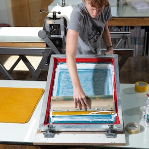 Peut inclure: Une personne utilise une raclette pour &eacute;taler de l'encre blanche sur un cadre de s&eacute;rigraphie. Le cadre est pos&eacute; sur une table avec un tissu jaune et un rouleau de ruban adh&eacute;sif.