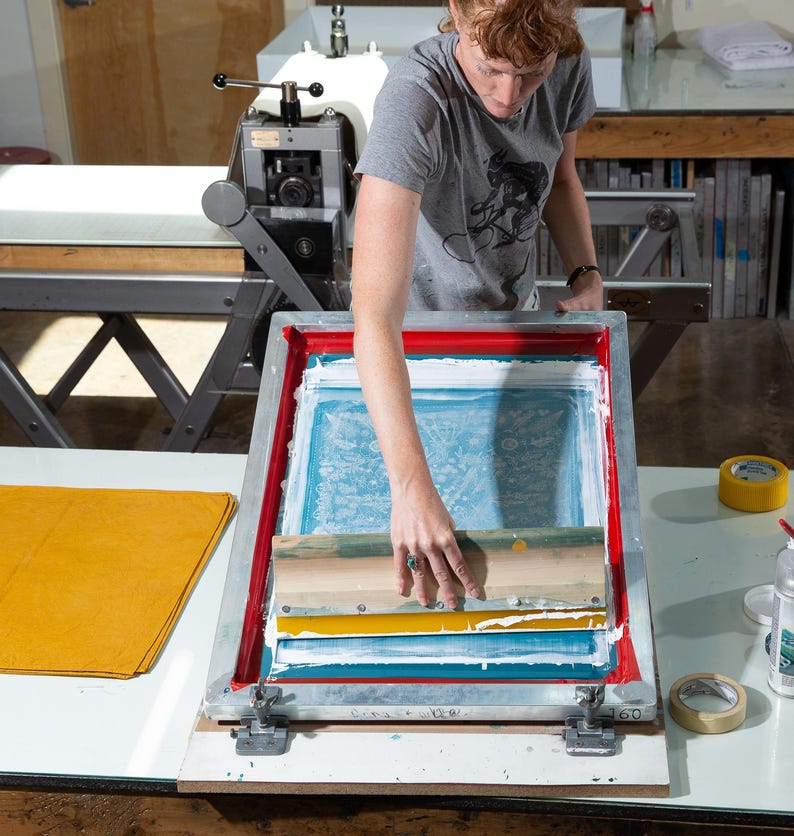 Peut inclure: Une personne utilise une raclette pour &eacute;taler de l'encre blanche sur un cadre de s&eacute;rigraphie. Le cadre est pos&eacute; sur une table avec un tissu jaune et un rouleau de ruban adh&eacute;sif.
