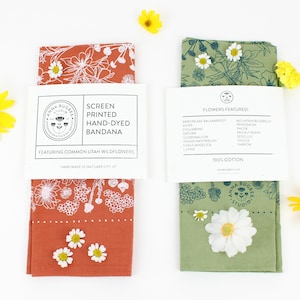 Peut inclure: Deux bandanas en coton teints &agrave; la main avec des illustrations de fleurs sauvages de l'Utah imprim&eacute;es &agrave; l'&eacute;cran. Le bandana de gauche est orange avec des fleurs blanches. Le bandana de droite est vert avec des fleurs blanches. Les bandanas sont 100% coton et faits &agrave; la main &agrave; Salt Lake City, Utah.