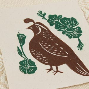 Op de afbeelding: Een vierkant stuk gebroken wit textiel met een bruine blokdruk van een kwartel die naar links kijkt, omringd door donkergroene florale elementen, waaronder twee kleine bloemen aan de linkerkant en een grotere groep van drie bloemen boven de vogel.