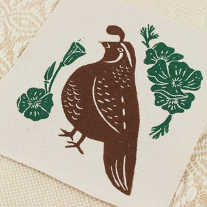 Op de afbeelding: Een vierkante, gebroken witte stoffen patch met een bruine blokdrukillustratie van een kwartel in profiel. De vogel wordt geflankeerd door twee groene botanische elementen, elk met een kleine bloem en bladeren, weergegeven in een eenvoudige grafische stijl.