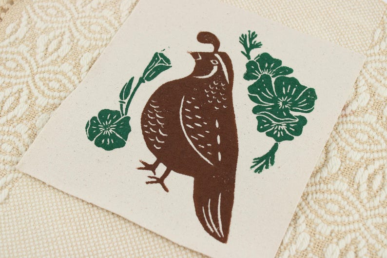 Op de afbeelding: Een vierkante, gebroken witte stoffen patch met een bruine blokdrukillustratie van een kwartel in profiel. De vogel wordt geflankeerd door twee groene botanische elementen, elk met een kleine bloem en bladeren, weergegeven in een eenvoudige grafische stijl.