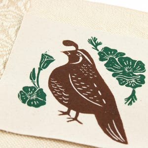 Op de afbeelding: Een vierkante, gebroken witte stoffen patch met een bruine blokdrukillustratie van een Californische kuifkwartel. De vogel wordt aan beide kanten geflankeerd door groene bloemmotieven. De patch ligt op een cr&egrave;mekleurige stof met een geweven patroon.