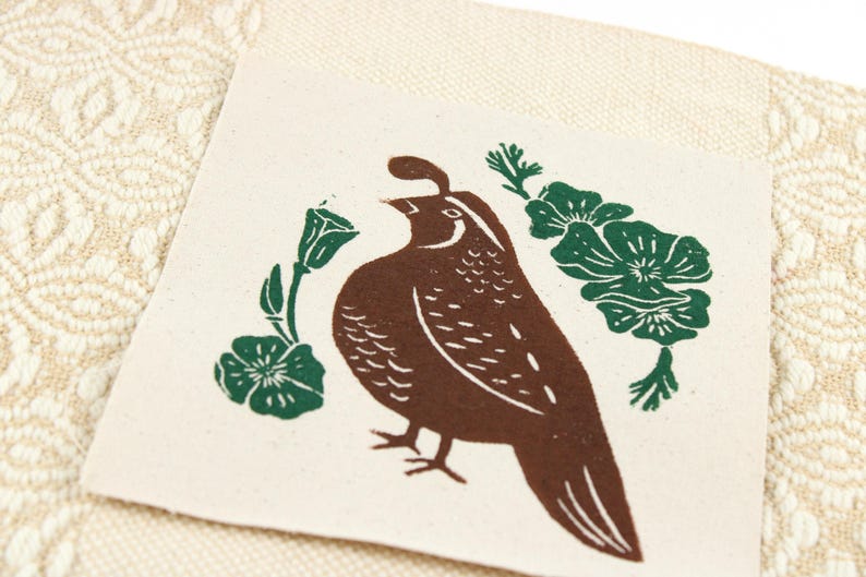 Op de afbeelding: Een vierkante, gebroken witte stoffen patch met een bruine blokdrukillustratie van een Californische kuifkwartel. De vogel wordt aan beide kanten geflankeerd door groene bloemmotieven. De patch ligt op een cr&egrave;mekleurige stof met een geweven patroon.
