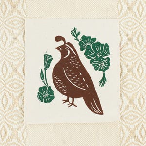 Op de afbeelding: Een vierkante stoffen patch met een bruine blokdrukillustratie van een kwartel, gecentreerd tussen groene bloemmotieven. Het ontwerp is gedrukt op gebroken wit canvas met een zichtbare weving en toont een vogel met een kuif.