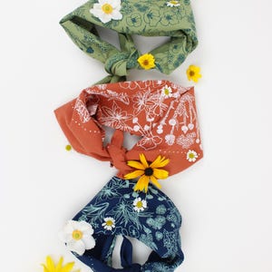 Peut inclure: Trois bandanas en tissu avec des motifs floraux. Les bandanas sont nou&eacute;s et dispos&eacute;s verticalement. Le bandana du haut est vert avec des fleurs blanches, le bandana du milieu est orange avec des fleurs blanches et le bandana du bas est bleu avec des fleurs blanches. Il y a des fleurs jaunes et blanches &eacute;parpill&eacute;es autour des bandanas.